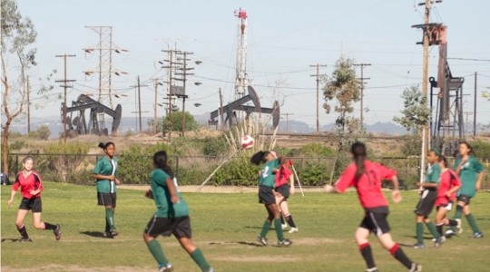 Young girls playing soccer under the shadow of a battery of oil derricks